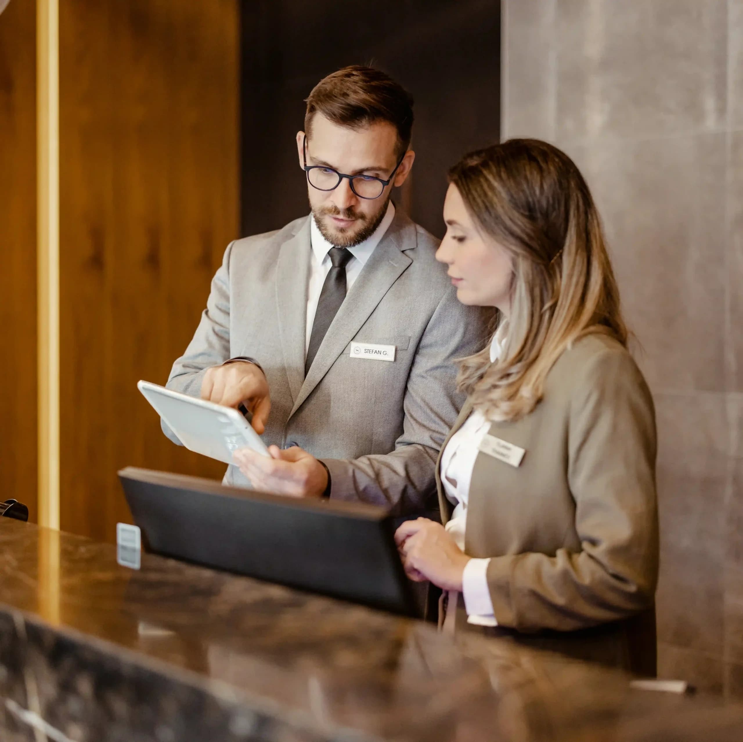 Two colleagues standing at a reception desk, reviewing how installing solar panels for their hotel has reduced energy costs.