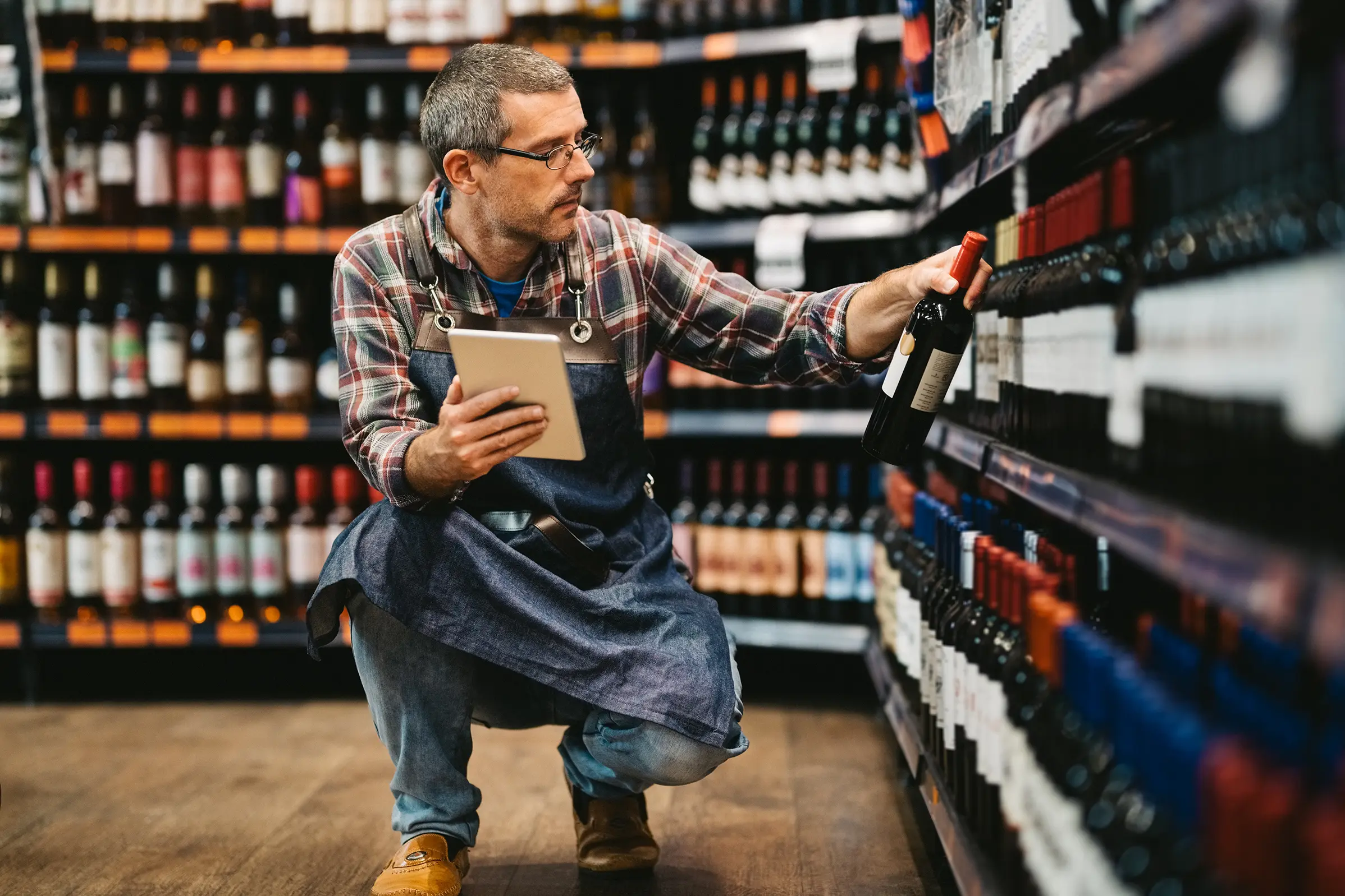 Supermarket employee in a large business stocking wine bottles on a shelf while using a tablet.
